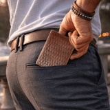 Lifestyle photo of a man in gray trousers tucking the cognac basketweave Mogul Billfold into his back pocket, leather belt and bracelets visible.