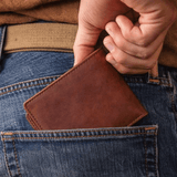Lifestyle photo of a cognac leather billfold wallet being tucked into the back pocket of blue jeans.