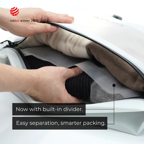 Hands lifting the grey divider inside a white Compak Large to separate black knitwear from cream and brown clothing, captioned Now with built-in divider, easy separation, smarter packing