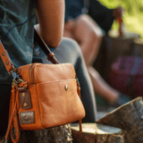 Close-up of the tan leather crossbody bag worn over the shoulder outdoors, showing the WILL Leather Goods patch and fringe tassel