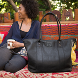 Lifestyle photo of a woman relaxing outdoors with the black leather tote resting beside her