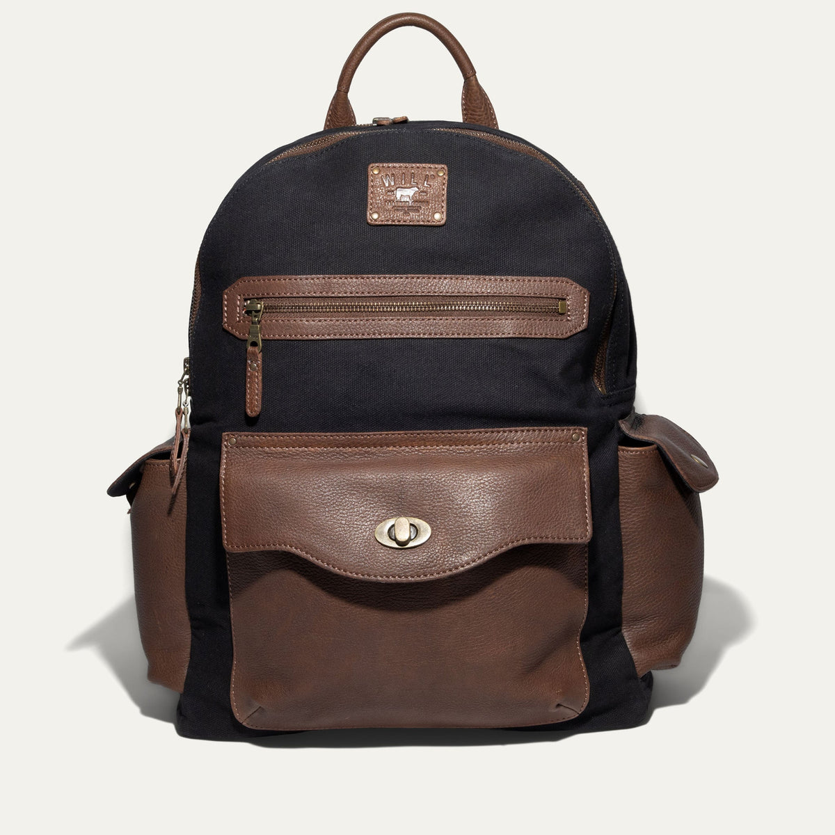 Will Leather Goods Canvas + Leather Backpack in black canvas with brown leather trim, front view showing the zip front pocket, flap pocket with turn-lock closure, and two side pockets, on a white background.