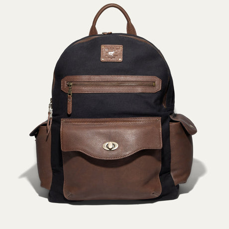 Will Leather Goods Canvas + Leather Backpack in black canvas with brown leather trim, front view showing the zip front pocket, flap pocket with turn-lock closure, and two side pockets, on a white background.