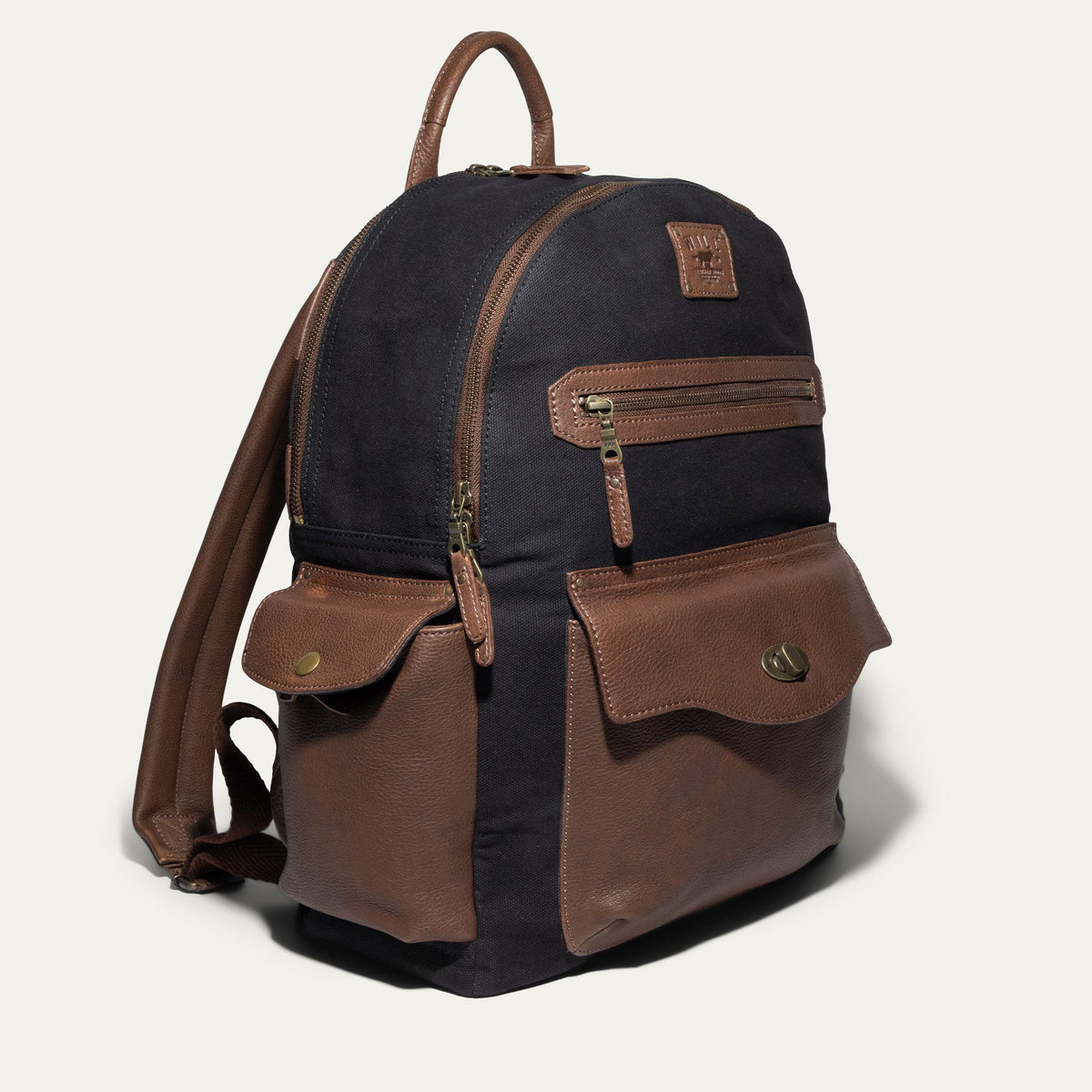 Side angle of the black canvas Canvas + Leather Backpack showing the brown leather top handle, side pockets, and adjustable straps.