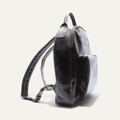 Side view of the dark brown leather backpack showing its rounded profile, front zip pocket, and adjustable shoulder strap.
