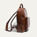 Side view of the tan leather backpack showing its rounded profile, front zip pocket, and side zipper.