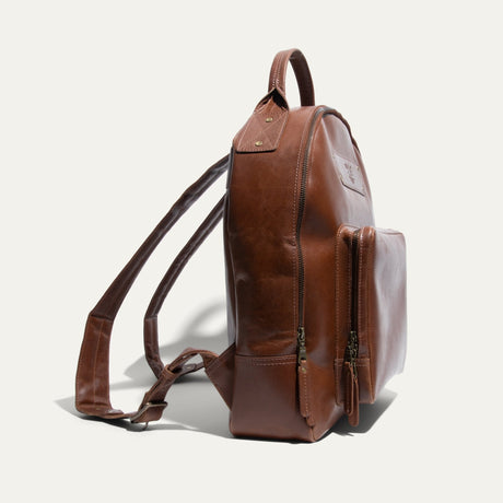 Side view of the tan leather backpack showing its rounded profile, front zip pocket, and side zipper.