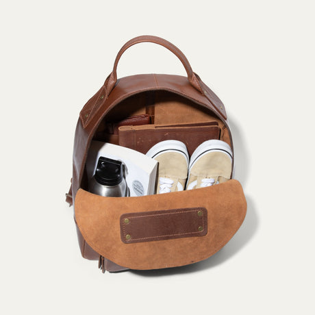 Tan leather backpack open and packed with a water bottle, book, wallet, and sneakers inside the main compartment.