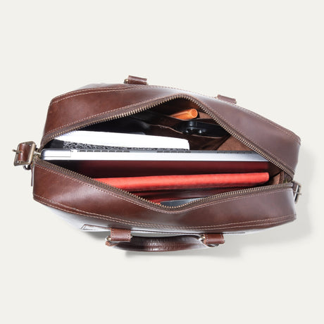 Top-down view of the dark brown leather briefcase unzipped, showing a laptop, notebook, pen, and phone inside