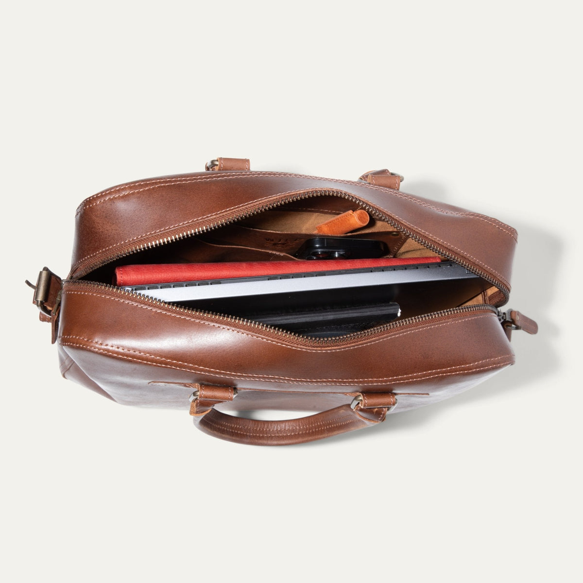 Top-down view of the tan leather briefcase unzipped, showing a laptop, tablet, phone, and red accessory pouch inside