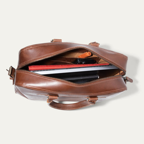 Top-down view of the tan leather briefcase unzipped, showing a laptop, tablet, phone, and red accessory pouch inside