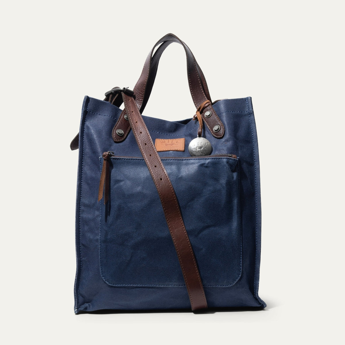 Navy waxed canvas market tote shown straight-on, with brown leather handles raised and an adjustable brown leather crossbody strap draped diagonally across the front zip pocket, plus a WILL leather tag and round metal fob