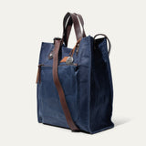 Navy waxed canvas market tote shown from a three-quarter angle, showing the side gusset, brown leather handles, and the crossbody strap's adjustable buckle