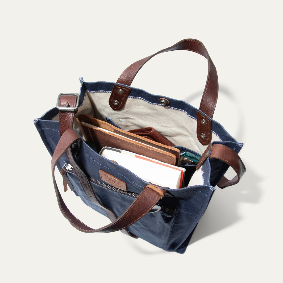 Overhead view looking down into the navy tote loaded with everyday items, including a wallet, a notebook, and a sunglasses case