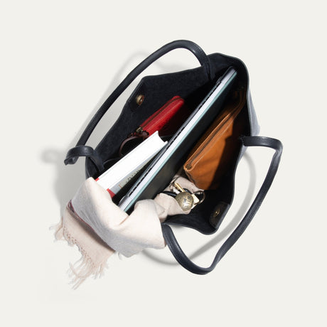 Overhead view into the open black Workshop Tote, showing the suede-lined interior packed with a laptop, notebooks, a red zip pouch, a tan zip pouch, keys, and a cream scarf.