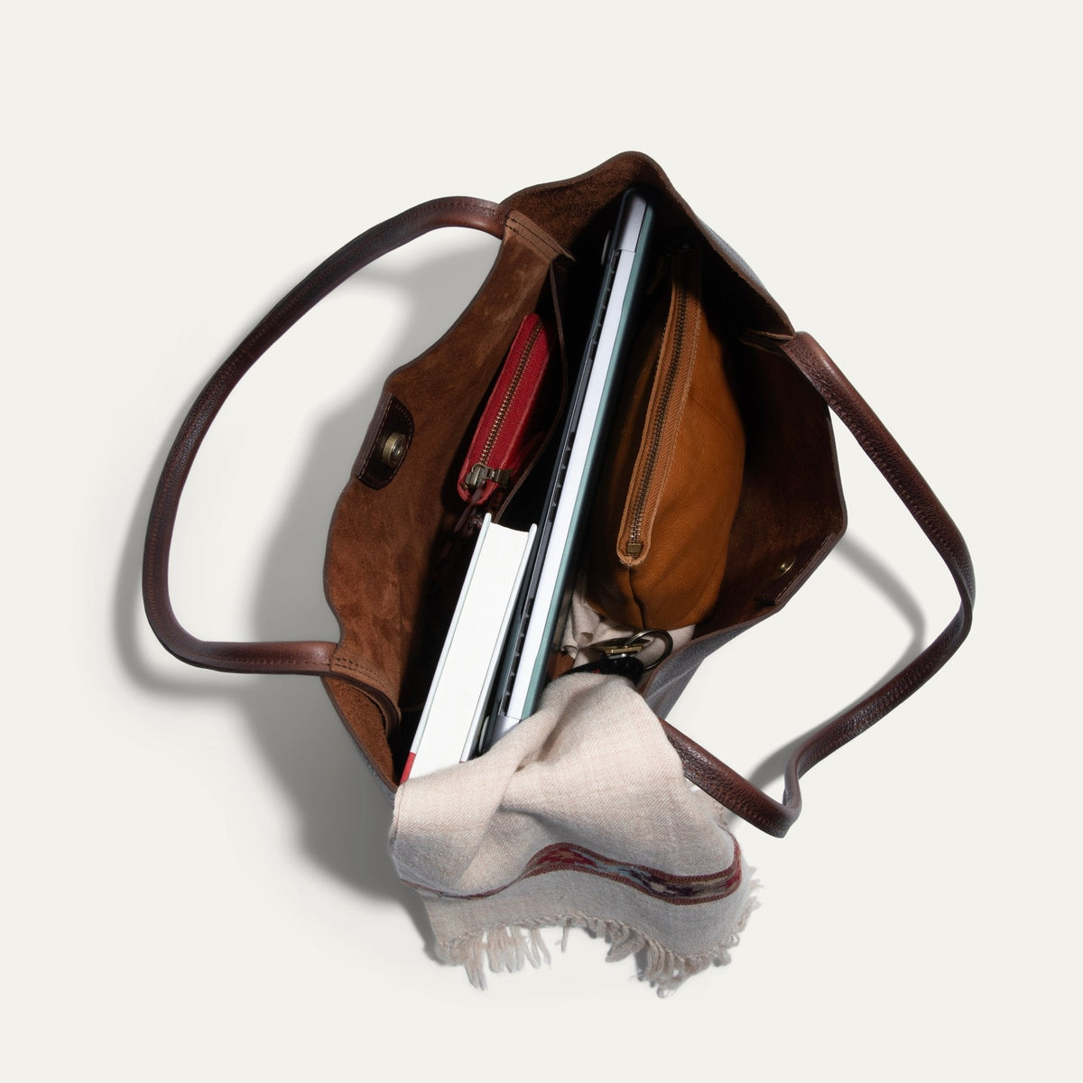 Overhead view into the open brown Workshop Tote, showing the suede-lined interior packed with a laptop, notebooks, a red zip pouch, a tan zip pouch, keys, and a cream scarf.