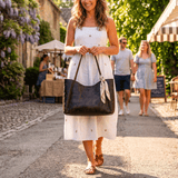 Lifestyle photo of a woman in a white sundress carrying the brown Workshop Tote by its straps while walking down a cobblestone street market.