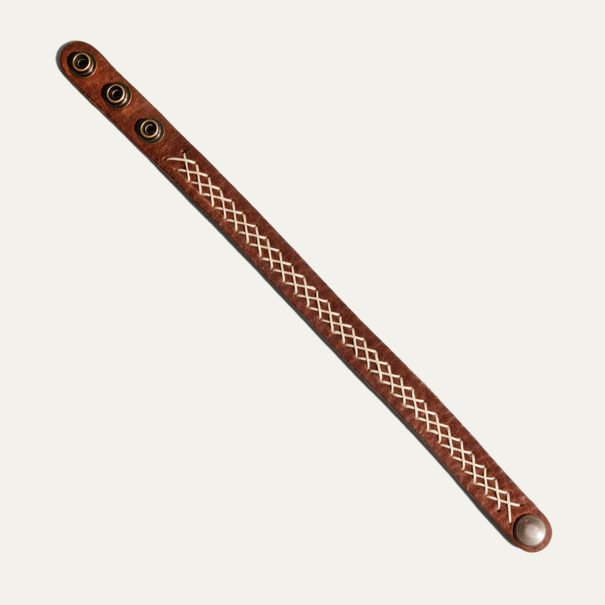 Flat diagonal view of the smooth grain side of the Cross Stitch Cuff, showing three brass snap studs at one end for size adjustment, a single line of cream stitching down the strap, and one snap stud at the opposite end.