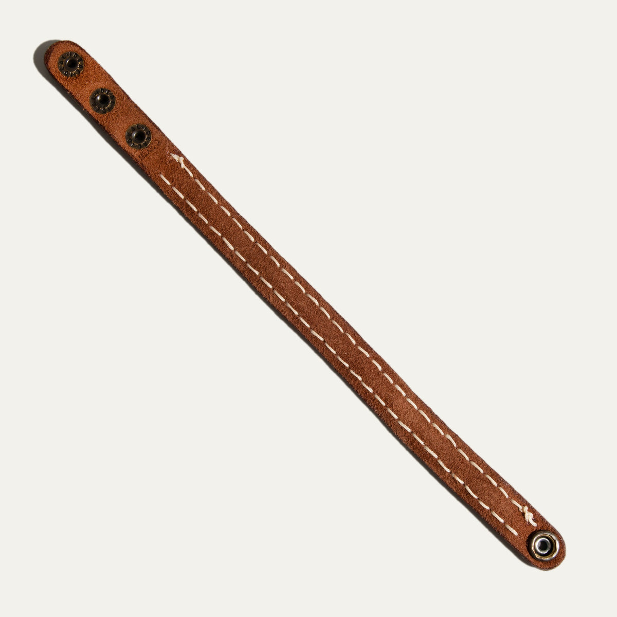 Flat diagonal view of the suede underside of the Cross Stitch Cuff, showing double rows of cream stitching down the strap, brass snap studs at one end and a metal eyelet at the other.