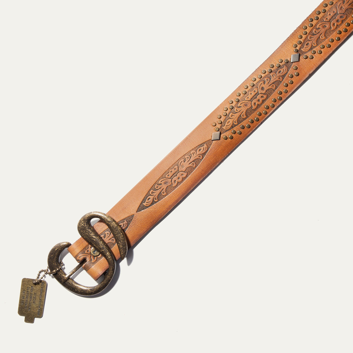 Close-up of the Baez Wild Ones Belt's antiqued brass double-prong buckle, tooled floral tabs, and brass stud trim on tan leather, with the Will Leather Goods buckle-change hang tag visible.