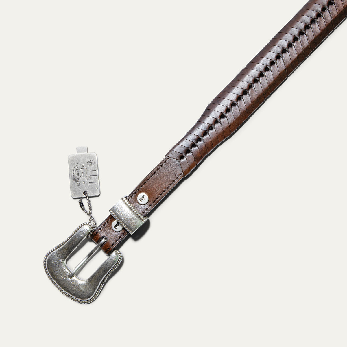 Close-up of the cognac brown tapered lace belt showing the woven lace-wrapped strap texture and silver keeper with a Will Leather Goods hang tag.