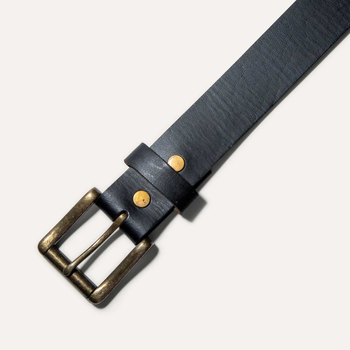 Close-up of the Black Edge Jean Belt buckle in black leather, showing the antique brass roller buckle and rivets on the keeper loop.