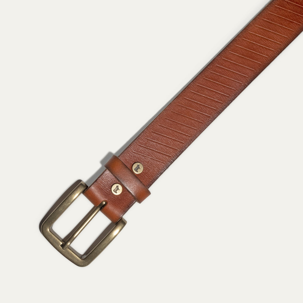 Close-up of the Razor Cut Belt buckle in cognac leather, showing the antique brass buckle, diagonal razor-cut striping, and embossed WILL Leather Goods dog logo snap.