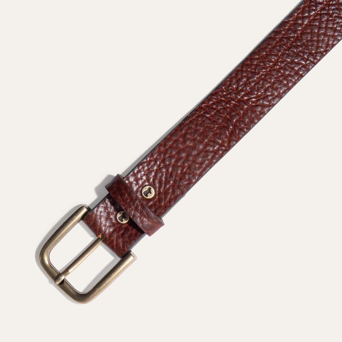 Close-up of the Shrunken American Bison Belt buckle in brown pebbled bison leather, showing the antique brass buckle and embossed WILL Leather Goods dog logo rivet.