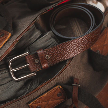 Lifestyle photo of a brown pebbled bison leather belt coiled inside an open canvas duffle bag beside leather luggage tags and gear.