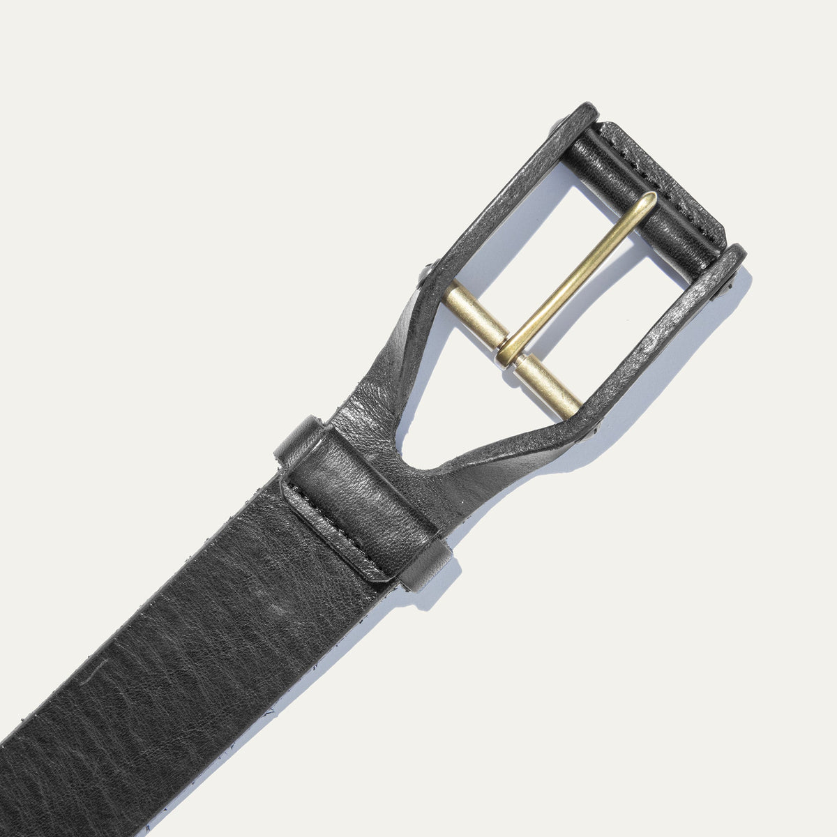 Close-up of the black leather buckle belt's brass roller buckle and keeper loop, on a white background.