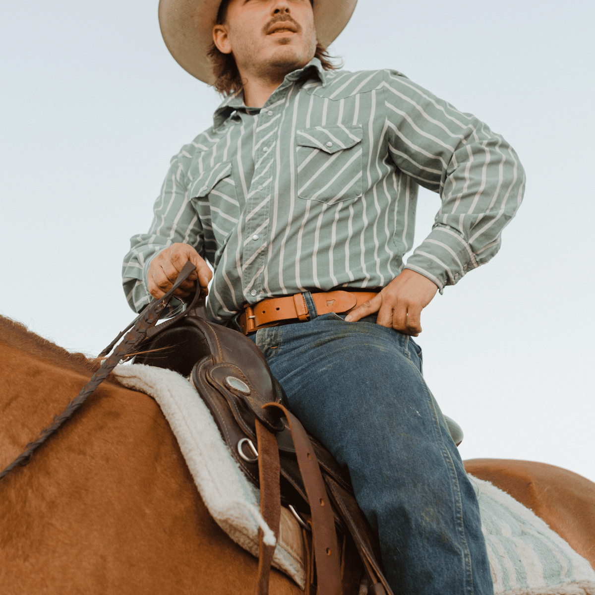 Lifestyle photo of a man on horseback in a striped western shirt and jeans, wearing the tan leather buckle belt with a cattle-brand buckle.