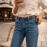 Lifestyle photo of a person wearing blue jeans with the black and tan Two-Tone Five Strand Braided Belt, standing at an outdoor Parisian cafe holding a coffee cup.