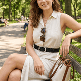 Lifestyle photo of a woman in a cream shirtdress seated on a park bench, wearing the black Twelve Strand Braided Belt with a brushed silver oval buckle.