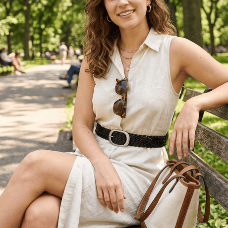 Lifestyle photo of a woman in a cream shirtdress seated on a park bench, wearing the black Twelve Strand Braided Belt with a brushed silver oval buckle.