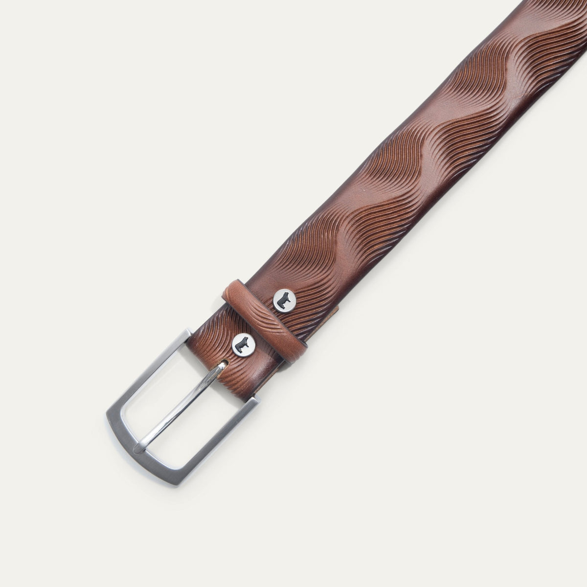 Close-up of the Will Leather Goods Wave Embossed Belt in cognac brown, showing the brushed silver buckle, keeper loop with embossed dog logo snap, and wave-textured leather.