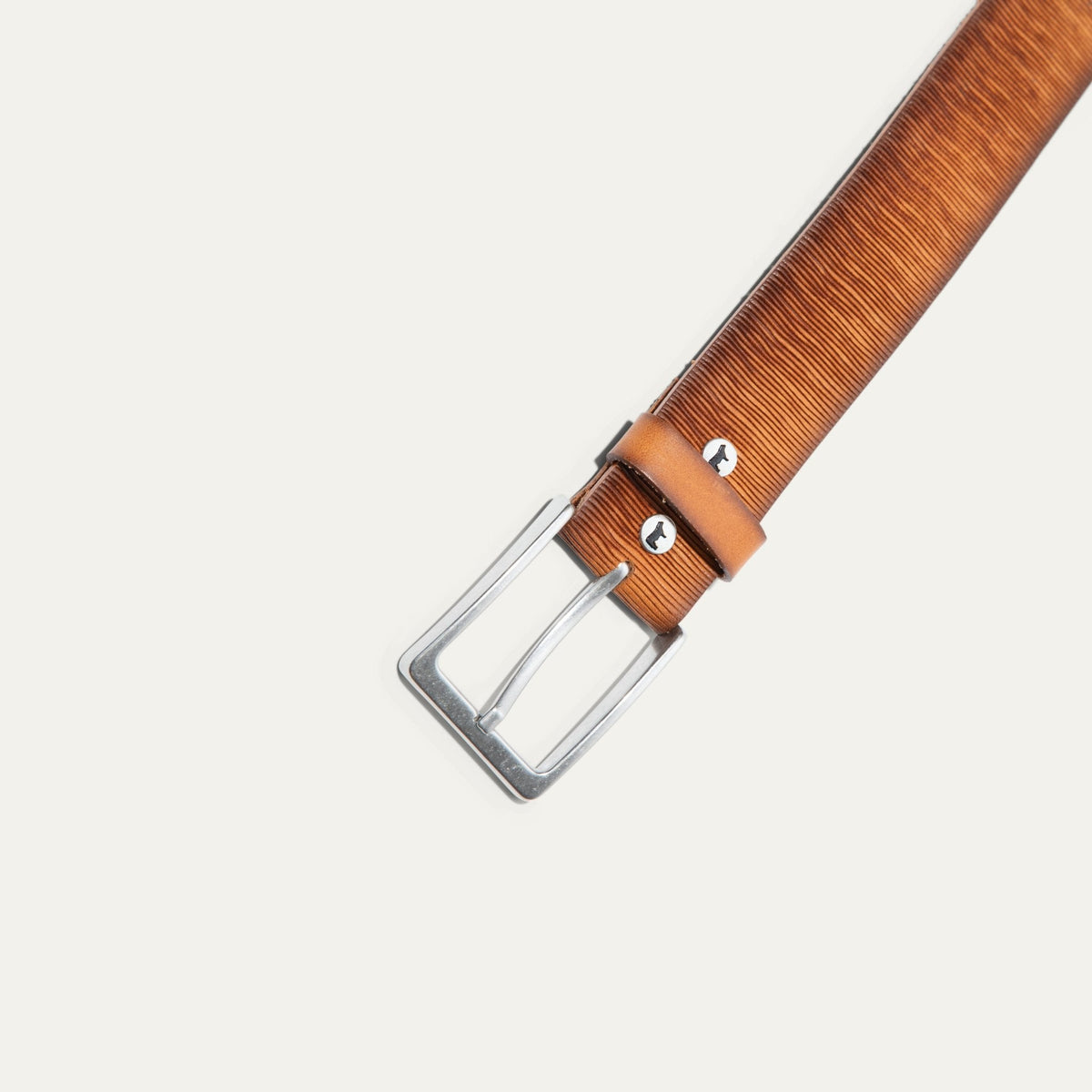 Close-up of the Wood Grain Embossed Belt's brushed silver buckle and keeper loop, showing the wood-grain embossed tan leather texture and a small Will Leather Goods dog logo stud.