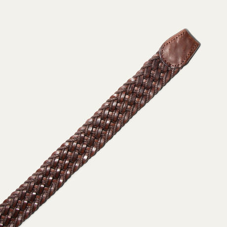 Close-up diagonal view of the dark brown braided belt's tip end, showing the woven leather and cord texture and stitched leather tab