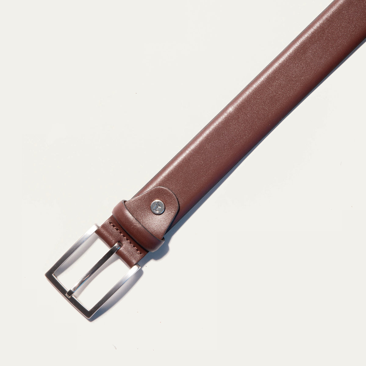 Close-up of the Will Leather Goods Glazed Calfskin Belt in cognac brown, showing the brushed silver buckle attachment, keeper loop, and rivet detail against a white background.