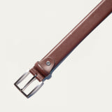 Close-up of the Will Leather Goods Glazed Calfskin Belt in cognac brown, showing the brushed silver buckle attachment, keeper loop, and rivet detail against a white background.