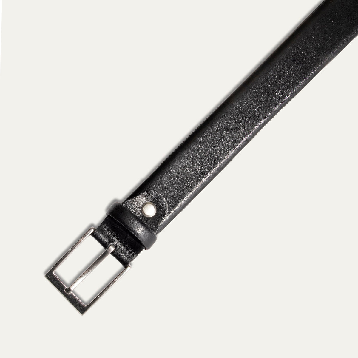 Close-up of the Will Leather Goods Glazed Calfskin Belt in black, showing the brushed silver buckle attachment, keeper loop, and rivet detail against a white background.