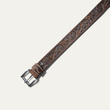 Close-up of the Hand-Tooled Morning Glory Jean Belt strap tip in antiqued brown leather, showing the hand-tooled floral scrollwork and row of adjustment holes.