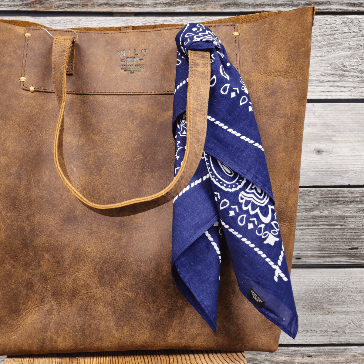 Lifestyle photo of the navy paisley Will's Bandana tied around the handle of a brown leather tote bag.
