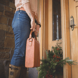 Lifestyle photo of a woman carrying the Will Leather Goods Hard Double Leather Wine Case in tan leather by the handle at a front doorway.