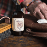 Lifestyle photo of a man's hands using a white cloth to apply Will Leather Goods All-In-One Leather Solution to a brown leather bag strap, bottle resting on the workbench beside it.