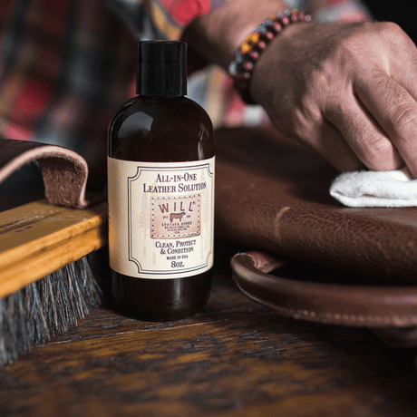 Lifestyle photo of a man's hands using a white cloth to apply Will Leather Goods All-In-One Leather Solution to a brown leather bag strap, bottle resting on the workbench beside it.