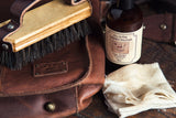 All-In-One Leather Solution bottle staged with a horsehair leather brush, a folded white cloth, and a brown leather bag stamped with the WILL leather patch.