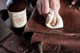 Close-up of a hand wiping All-In-One Leather Solution onto a brown leather bag flap with a white cotton cloth, showing brass hardware and stitched seams.