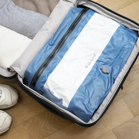 A Steel Blue TILLIV Compak Large packed with clothing in an open suitcase, white trainers on the floor beside it