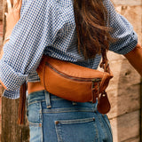 Lifestyle photo of a woman wearing the tan leather fanny pack across her back with a gingham shirt and jeans