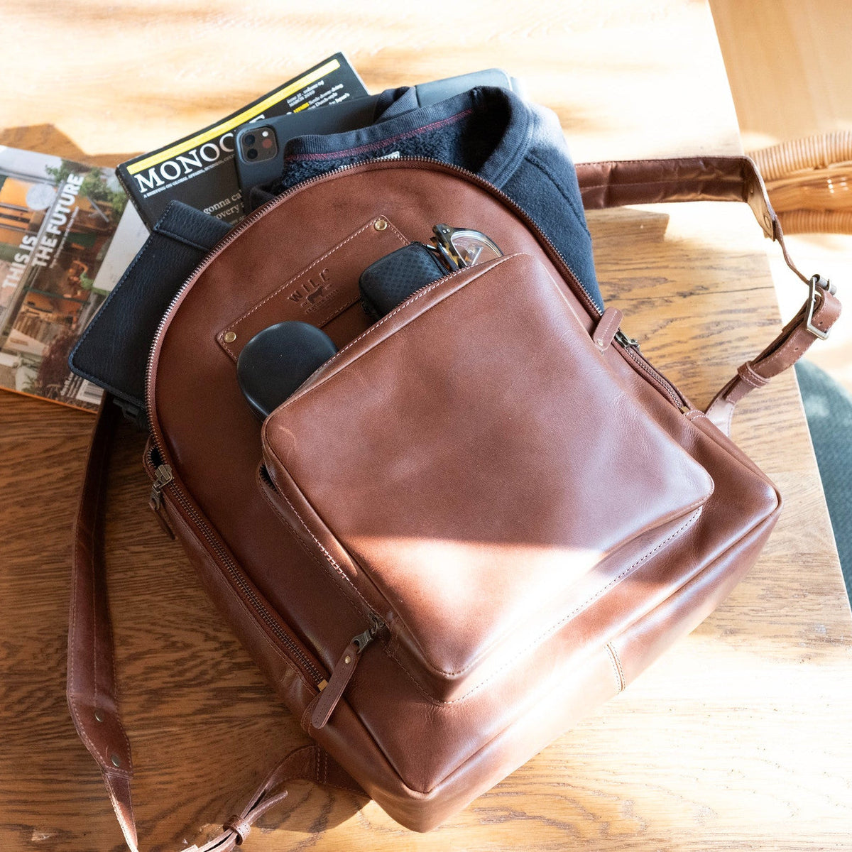 Tan leather backpack open and packed on a wood table with a magazine, phone, sunglasses, and other items visible inside.